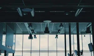 Synliga ventilationsrör och belysning i taket, med stora fönster som visar en molnig himmel och en grönskande kulle.