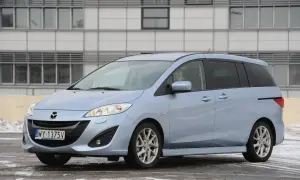 Jasnoniebieska Mazda 5 z nowymi żarówkami w reflektorach, zaparkowana przed nowoczesnym budynkiem.