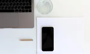 Przygotowanie do naklejania szkła na telefon. Obok laptopa i szklanki wody leży czarny smartfon na białej kartce.