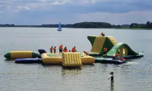 Rodzinna zabawa na wodnym placu zabaw na Mazurach. Dzieci w kamizelkach ratunkowych wspinają się i ślizgają po dmuchanych konstrukcjach.