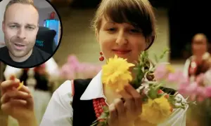 Mężczyzna w okrągłym kadrze mówi, podczas gdy młoda kobieta w tradycyjnym stroju, z koralami i kolczykami, trzyma bukiet żółtych kwiatów.