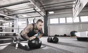 Mann mit Tattoos macht Liegestütze auf einem Medizinball, fokussiert auf sein hypertrophie training. Im Hintergrund ein Boxring und Trainingsgeräte.