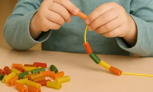 Manos de niño ensartando macarrones de colores, desarrollando la escritura manual.