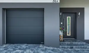 Nowoczesne drzwi wejściowe z wąskim oknem, obok nich szara brama garażowa.