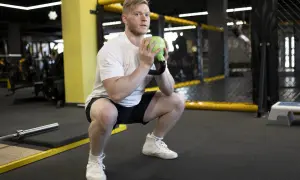Mężczyzna wykonuje goblet squats z zielonym kettlebellem w siłowni.
