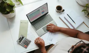 Biurko z laptopem, kalkulatorem, dokumentami i kawą, osoba pracuje nad finansami.
