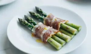 Zielone szparagi owinięte w cienkie plastry szynki parmeńskiej, polane oliwą, na białym talerzu.