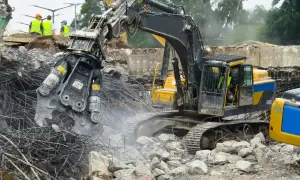 Maszyna budowlana wykonuje rozbiórkę budynku, krusząc beton i stal. W tle widać pracowników w kamizelkach odblaskowych.