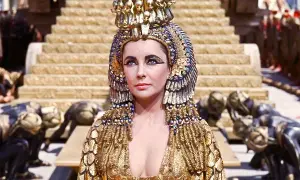 Elizabeth Taylor jako Kleopatra w złotej, bogato zdobionej sukni i koronie, z poddanymi klęczącymi u stóp.