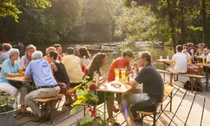 Menschen genießen das Leben im cafe am neuen see. Sie sitzen an Holztischen, trinken Bier und unterhalten sich. Im Hintergrund fließt ein ruhiger Fluss.