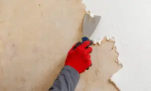 Hand med röd handske skrapar bort gammal strukturtapet med en spackel.