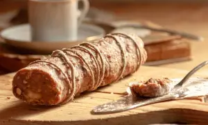 Ein rustikaler salame al cioccolato, gebunden mit Juteschnur, liegt auf einem Holzbrett. Daneben eine Kaffeetasse und ein Stück auf einem Tortenheber.
