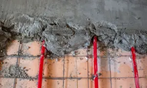 Budowa podłogi z ogrzewaniem podłogowym. Widać siatkę zbrojeniową, czerwone rurki i świeżą wylewkę cementową. Jaki cement na wylewki będzie najlepszy?