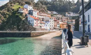 Una mujer admira la costa de **pueblos de Asturias cerca de Galicia**. Casas coloridas se aferran a la ladera verde junto a un mar turquesa.