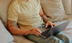 Man med tatuering på armen sitter i soffan med laptop, kanske för att han är hemma under en ny sjukperiod.