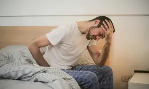 Man met hand op hoofd, zittend op bed, worstelend met hoe in slaap vallen met stress.