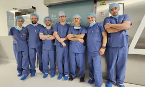 Grupa lekarzy w niebieskich uniformach i czepkach stoi przed drzwiami sali operacyjnej.