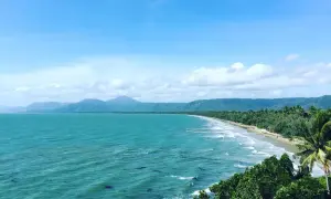 Vacker kustlinje vid Port Douglas med turkost hav, grönskande berg och en gyllene strand under en molnig himmel.