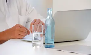 Lekarz w białym kitlu przy laptopie, obok szklanka wody i butelka. Czy można pić wodę przed tomografią z kontrastem? Zapytaj lekarza.