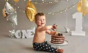 Uśmiechnięty chłopiec z czekoladą na rękach świętuje z okazji pierwszych urodzin. Przed nim tort, a w tle balony i cyfra 1.