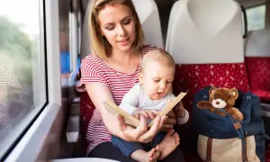 Mama czyta książeczkę rocznemu dziecku w pociągu. Obok siedzi plecak z pluszowym misiem.