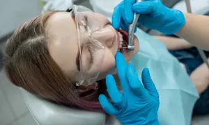 Dentysta w niebieskich rękawiczkach wykonuje zabieg, który wyjaśnia, co to jest leczenie kanałowe. Pacjentka ma okulary ochronne.