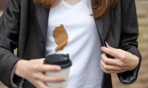 Frau mit Kaffeefleck auf weißem T-Shirt unter dunklem Blazer. Ein Becher Kaffee in der Hand. Tipps, wie man kaffeeflecken aus kleidung entfernen kann.