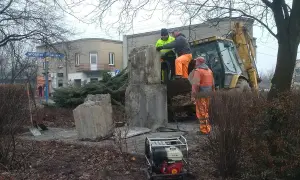 Prace przy pomniku, gdzie widać objawy zapadania się ziemi. Pracownicy w pomarańczowych strojach i koparka próbują wzmocnić podłoże.