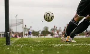 Piłkarz kopie piłkę podczas sparingów. W tle widać innych zawodników i bramkę.