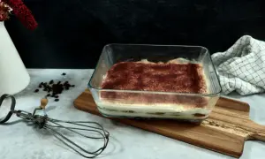 Un délicieux tiramisu 6 personnes, prêt à être dégusté, saupoudré de cacao. Un fouet ancien et des grains de café complètent la scène.