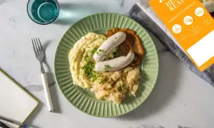 Fertig zubereitete Weißwurst mit Kartoffelpüree und Sauerkraut auf einem grünen Teller. Anleitung zum Weißwurst kochen liegt daneben.
