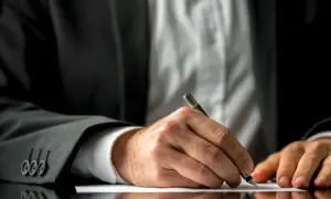 Mężczyzna w garniturze pisze piórem na białej kartce papieru.