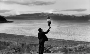 Un homme soulève un bébé au-dessus de l'eau, une scène qui semble être une mise en scène poétique avec des montagnes en arrière-plan.