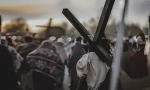 Osoba z koroną cierniową niesie krzyż, idąc w procesji. W tle widać inne krzyże i ludzi, przypominające stacje drogi krzyżowej.