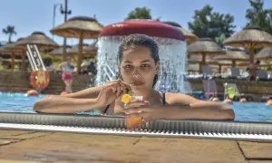 Kobieta z drinkiem w basenie, w tle wodospad i parasole. To kwintesencja wakacji all inclusive plus co to znaczy – beztroska i relaks.