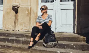 Elegancka kobieta w czarnych cygaretkach i bluzce w paski, siedząca na schodach. Stylowa torebka Michael Kors dopełnia całość.