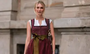 Blonde Frau in einem roten Dirndl mit weißer Bluse und einer grünen Schürze. Sie trägt eine Dior-Tasche.