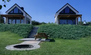 Dwa nowoczesne domki na plaży Bałtyk, z tarasami i dużymi oknami, otoczone zielenią. Obok schody, ławka piknikowa i palenisko.