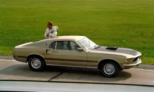 En man med hjälm lutar sig mot en guldig mustang 60 tal på en bana.