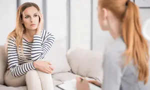 Kobieta w paski siedzi na kanapie, zamyślona, podczas rozmowy z terapeutką. Czy psycholog pomaga? Tak, widać, że wsparcie jest potrzebne.