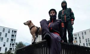 Zwei Personen in Gorpcore-Outfits und ein Hund stehen auf einem Metallgitter vor modernen Gebäuden.