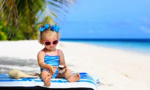 Mała dziewczynka w okularach przeciwsłonecznych i stroju kąpielowym bawi się muszlami na plaży. To idealne wakacje z niemowlakiem!