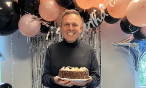 Uśmiechnięty mężczyzna trzyma tort na przyjęciu z balonami i błyszczącą kurtyną. To idealna niespodzianka na urodziny dla faceta!