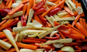 En ugnsplåt fylld med färgglada rotfrukter i ugn: morötter, palsternacka, paprika och lök, kryddade med örter.