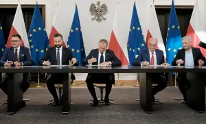 Pięciu mężczyzn w garniturach siedzi przy stole, podpisując dokumenty. W tle flagi Polski i UE. Może to być ważny moment dla sejmowej komisji śledczej.
