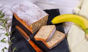 Ein saftiger Bananenkuchen, bestäubt mit Puderzucker, liegt auf einem schwarzen Brett. Daneben zwei Scheiben und eine reife Banane. Das ist der beste Bananenkuchen der Welt!