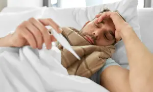 Chory mężczyzna z termometrem w ręku, owinięty w koc, czuje się źle. Domilgan.