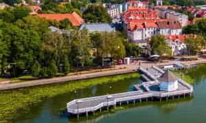Ostróda atrakcje: malowniczy deptak nad jeziorem z białym molo i altanką, w tle zabudowa miejska i zieleń.