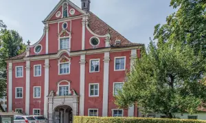 Prachtvolles Gebäude des Klosters Salem mit roter Fassade, weißen Säulen und einem geschwungenen Giebel. Ein Baum spendet Schatten.