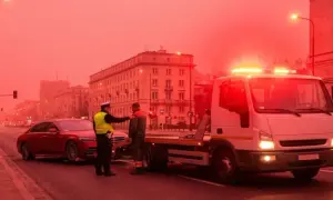 Policjant nadzoruje konfiskatę samochodu. Czerwony sedan jest ładowany na lawetę, podczas gdy migające światła ostrzegawcze oświetlają scenę w mieście.